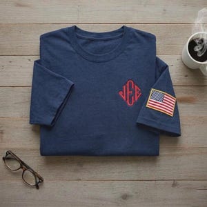 May include: Navy blue t-shirt with a red embroidered monogram and an American flag patch on the sleeve. The shirt is folded on a wooden surface, with a cup of coffee and eyeglasses nearby.