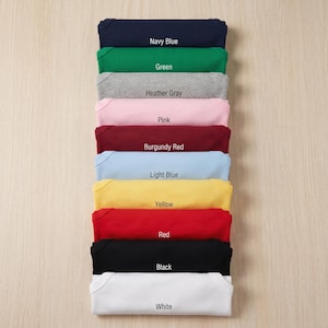 May include: A stack of folded baby onesies in various colors, including navy blue, green, heather gray, pink, burgundy red, light blue, yellow, red, black, and white. Each onesie has a label with its color name.