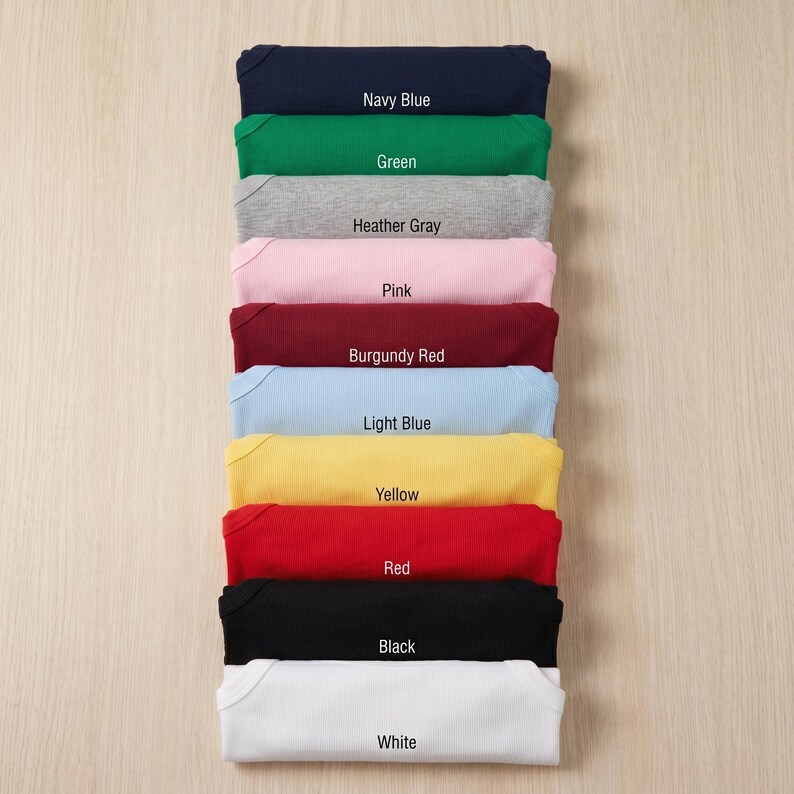 May include: A stack of folded baby onesies in various colors, including navy blue, green, heather gray, pink, burgundy red, light blue, yellow, red, black, and white. Each onesie has a label with its color name.