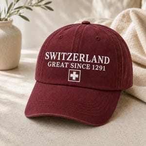 Op de afbeelding: Een kastanjebruine baseballpet met witte geborduurde tekst SWITZERLAND GREAT SINCE 1291 boven een wit vierkant met een wit Zwitsers kruis. De pet heeft een gebogen klep en een gestructureerde kroon met zichtbare stiksels.