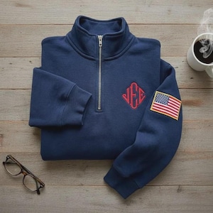 Può includere: Felpa blu navy con zip a un quarto, monogramma rosso e toppa con bandiera americana sulla manica. La felpa è piegata su una superficie di legno, con una tazza di caffè e occhiali nelle vicinanze.