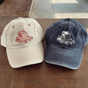 May include: Two baseball caps, one beige and one navy blue, are displayed. Each cap features a graphic of a melting ice cube. The beige cap has a red graphic, while the navy cap has a white graphic. Both caps have a curved brim.