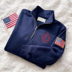 May include: Navy blue quarter-zip sweatshirt with a red embroidered logo and an American flag patch on the sleeve. A small American flag is placed next to the sweatshirt. The sweatshirt has a ribbed cuff and collar.
