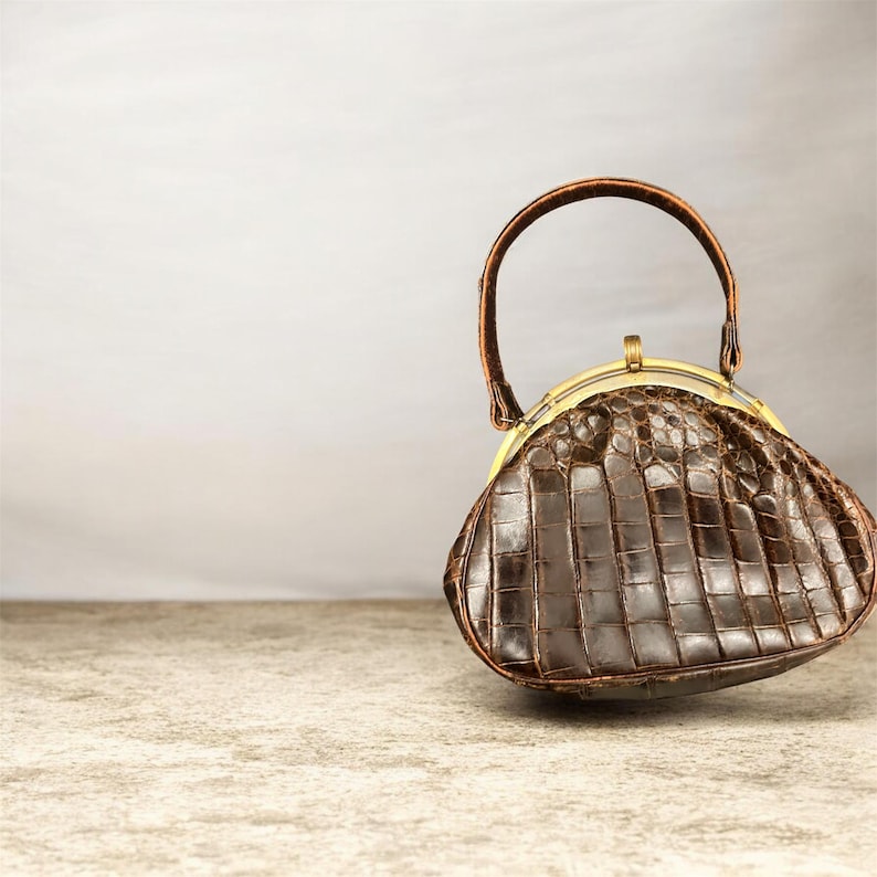 K&ouml;nnte beinhalten: Eine vintage braune Handtasche mit strukturierter, krokodil&auml;hnlicher Textur. Die Tasche hat einen geschwungenen Griff und einen goldfarbenen Verschluss. Die Tasche ist vor einem neutralen Hintergrund platziert, was ihr klassisches Design hervorhebt.