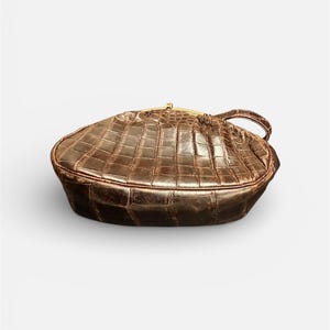 K&ouml;nnte beinhalten: Braune, strukturierte Handtasche mit goldfarbenem Verschluss und Griff. Die Tasche hat eine rechteckige Form mit abgerundeten Kanten und einem krokodilartigen Muster. Der Griff ist klein und an der Seite der Tasche befestigt.