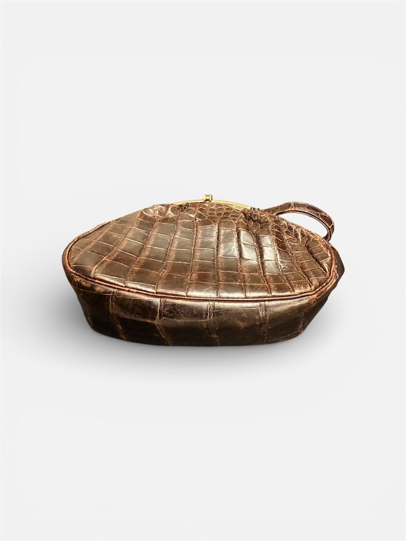 K&ouml;nnte beinhalten: Braune, strukturierte Handtasche mit goldfarbenem Verschluss und Griff. Die Tasche hat eine rechteckige Form mit abgerundeten Kanten und einem krokodilartigen Muster. Der Griff ist klein und an der Seite der Tasche befestigt.
