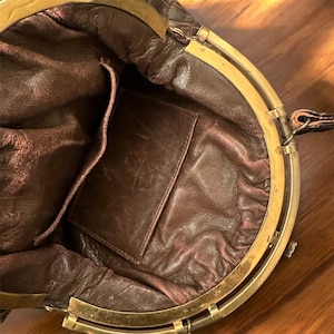 K&ouml;nnte beinhalten: Offene Ansicht einer Vintage-Handtasche aus braunem Leder. Der Innenraum hat ein dunkelbraunes Futter und eine kleine Tasche. Die Tasche hat einen goldfarbenen Metallrahmen und -verschluss. Das Leder weist Gebrauchsspuren auf, was ihren Vintage-Charme unterstreicht.