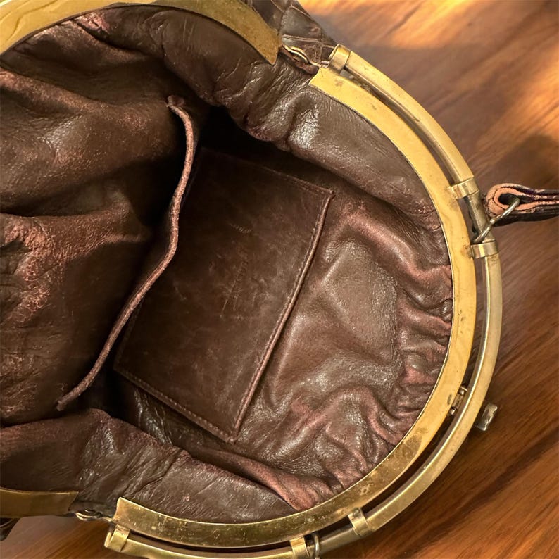 K&ouml;nnte beinhalten: Offene Ansicht einer Vintage-Handtasche aus braunem Leder. Der Innenraum hat ein dunkelbraunes Futter und eine kleine Tasche. Die Tasche hat einen goldfarbenen Metallrahmen und -verschluss. Das Leder weist Gebrauchsspuren auf, was ihren Vintage-Charme unterstreicht.