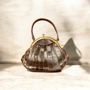 K&ouml;nnte beinhalten: Eine vintage braune Handtasche mit einem strukturierten, krokodilartigen Muster. Die Tasche hat einen gebogenen Griff und einen goldfarbenen Verschluss. Das Design der Tasche erinnert an klassische Stile.