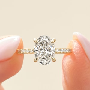May include: A gold engagement ring with an oval-cut diamond center stone. The band is adorned with small, sparkling diamonds. The ring is held between two fingers against a soft, neutral background.