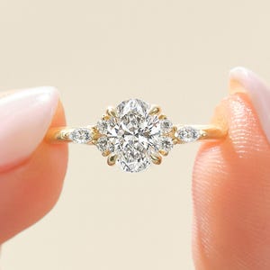 Może przedstawiać: Złoty pierścionek zaręczynowy z centralnym diamentem o owalnym szlifie, otoczonym mniejszymi diamentami. Pierścionek ma również małe diamenty na obrączce. Pierścionek jest trzymany między dwoma palcami.