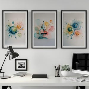 May include: Three framed watercolour prints. The prints feature abstract designs with splashes of blue, yellow, and orange. One print depicts a microscope. The frames are black, and the prints are displayed on a white desk with a black lamp and computer.
