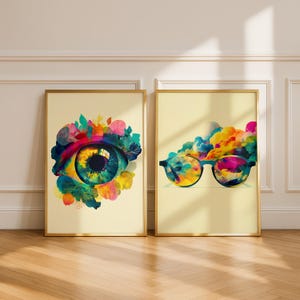 May include: Two framed art prints. One features a colorful eye with floral accents, the other a pair of glasses with a rainbow cloud design. Both prints have gold frames and are set against a cream background.