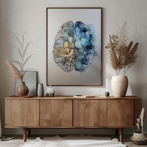 Op de afbeelding: Een ingelijste muurkunstprint met een illustratie van een gespleten brein. De linkerkant toont ingewikkeld zwart lijnwerk met goudgele aquarelaccenten, terwijl de rechterkant vloeiende, abstracte blauwe en marineblauwe aquarelkleuren bevat.
