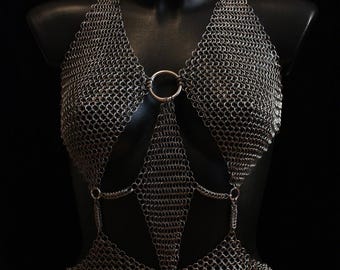 Könnte beinhalten: Ein silberfarbenes Kettenhemd-Haltertop mit geometrischem Design. Das Top hat einen Neckholder, einen zentralen Ring und rautenförmige Paneele, die durch kleinere Ringe verbunden sind. Das Kleidungsstück wird auf einer schwarzen Schaufensterpuppe vor schwarzem Hintergrund präsentiert.
