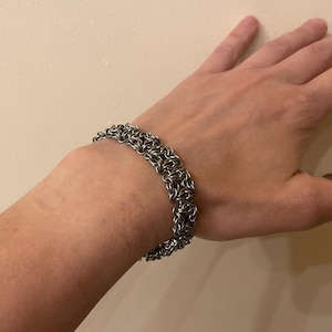 Puede incluir: Un brazalete de cota de malla de color plateado. El brazalete está hecho de pequeños anillos de metal entrelazados, creando una banda texturizada y flexible. El brazalete se usa en la muñeca, mostrando su intrincado diseño y artesanía.