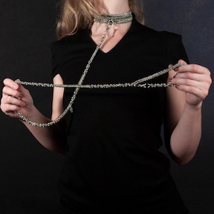 Puede incluir: Collar gargantilla de cadena plateada con una cadena para el cuerpo a juego. La gargantilla presenta un diseño de múltiples hilos, y la cadena para el cuerpo se extiende hacia abajo, sostenida por la persona. La joyería se muestra sobre un fondo oscuro.