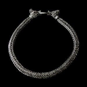 May include: A silver-toned necklace with a woven chain design. The necklace features two dragon head terminals at the clasp. The background is solid black, highlighting the intricate details of the jewelry.
