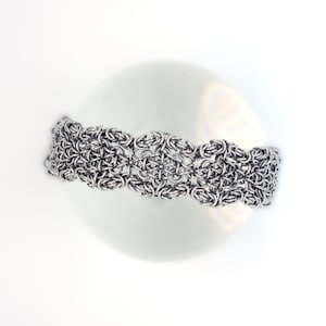 May include: A silver-toned bracelet with a detailed chainmail design. The bracelet is composed of interconnected metal rings, creating a textured, woven appearance. The intricate pattern is centered on a white, spherical object, creating a striking contrast.