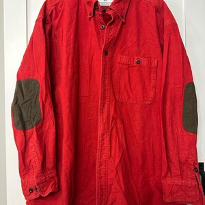 May include: A bright red button-down shirt with long sleeves and a chest pocket. The shirt features brown elbow patches and a button-down collar. The shirt is hanging on a black hanger against a white door.