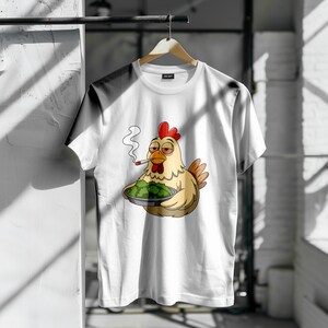May include: White t-shirt with a cartoon chicken smoking a cigarette and holding a plate of green food. The chicken has a red comb. The shirt is displayed on a wooden hanger.