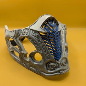 Puede incluir: Una máscara plateada y azul con detalles intrincados. La máscara presenta una sección de malla azul y patrones ornamentados. El fondo es amarillo.