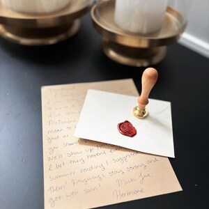 May include: A white envelope with a red wax seal and a wooden stamp, resting on a brown paper with handwritten text. Two gold candle holders with white candles are in the background.