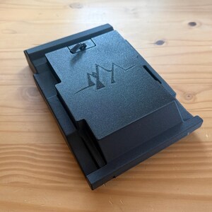 May include: A black, rectangular device with a mountain-like logo on the top surface. The device has a hinged top and a textured finish. It is resting on a wooden surface.