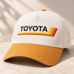 Peut inclure: Une casquette de baseball couleur crème avec une visière jaune moutarde et un petit bouton assorti sur le dessus. L'avant présente le texte brodé noir TOYOTA au-dessus d'un graphique horizontal composé de trois bandes parallèles jaune, orange et rouge.