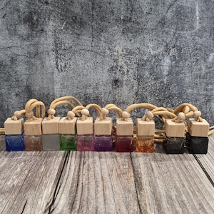 May include: A collection of small glass bottles with wooden lids and beige cords. The bottles contain liquids in various colours, including blue, orange, and green. The background is a textured grey wall and a wooden surface.