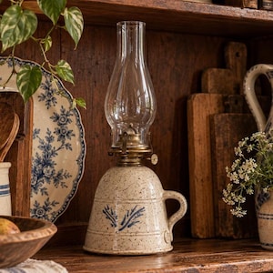 May include: A ceramic oil lamp with a speckled cream finish and a blue painted leaf design. The lamp features a brass burner mechanism and a clear glass chimney. It has a rounded base with a side handle, providing a stable structure for the light.