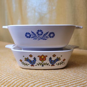 May include: Two white, rectangular casserole dishes stacked. The top dish has a blue floral design. The bottom dish features blue birds, orange flowers, and green foliage. Both dishes have handles for easy use.