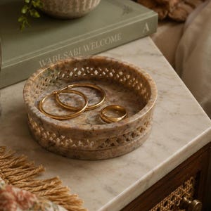 May include: A round, beige, speckled stone tray with a decorative lattice-cut side wall. Inside the tray are two gold hoop earrings and one plain gold band ring. The stone has a polished finish with natural variations in colour and texture.
