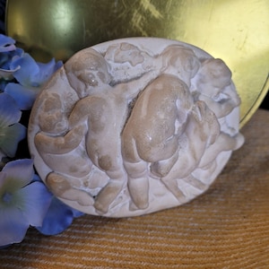 May include: A circular, off-white bas-relief sculpture depicting several cherubic figures in a playful scene. The figures are rendered in a classical style, with soft, rounded forms and subtle shading. The sculpture is set against a blurred background.