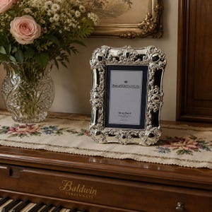 Peut inclure: Cadre photo argenté avec des détails ornés. Le cadre est rectangulaire avec un intérieur noir et le texte "PHILIP WHITNEY LTD" et "Silver-Plated". Il est posé sur un piano avec un chemin de table floral et un vase de fleurs.