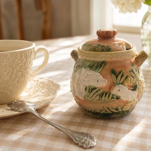 Peut inclure: Un sucrier en céramique avec couvercle, orné de motifs en relief représentant des lapins blancs et du feuillage vert sur un fond pêche. Le récipient possède deux petites anses et un bord texturé. Une tasse crème et une cuillère sont présentes.