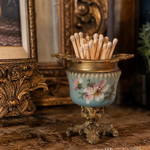 May include: A decorative match holder featuring a light blue glass body with hand-painted pink floral motifs, set upon an ornate gold-toned metal pedestal base with matching gold-toned metal rim and handles, filled with wooden matches.