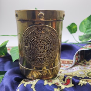 Peut inclure: Un récipient cylindrique en laiton avec un motif de calendrier aztèque détaillé. Le récipient a une finition polie et des rivets sur le dessus. Le fond présente un tissu bleu et de la verdure. Environ 10 cm de haut.