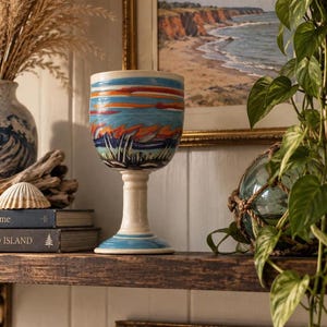 May include: A ceramic goblet featuring a hand-painted landscape design with horizontal bands of blue, orange and white. The vessel has a wide bowl, a slender stem and a circular base, all finished with a glossy glaze over the colourful artwork.