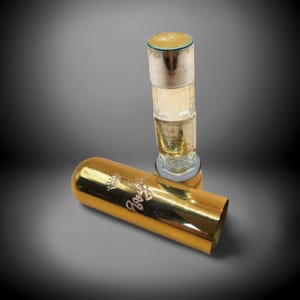 May include: A gold-coloured perfume bottle with a matching cylindrical cap. The bottle is clear with a gold-coloured top and base. The cap is lying on its side, displaying the word "Goya" in cursive script.