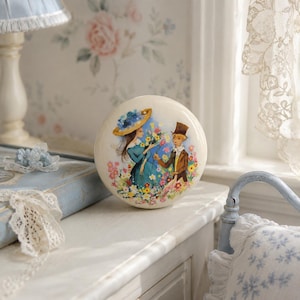 May include: A circular, cream-coloured ceramic knob with a vintage illustration of a woman in a blue dress and hat and a man in a brown suit, surrounded by colourful flowers. The knob is likely for a drawer or cabinet, adding a touch of vintage charm.