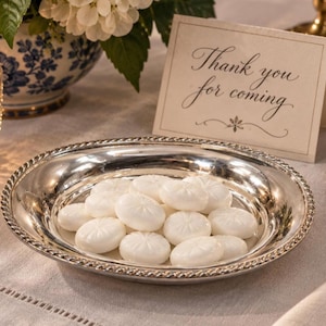 May include: A round silver tray with a twisted rope-style rim containing several small, round, white sweets with a starburst pattern on top. Behind the tray sits a cream-coloured folded card with the text Thank you for coming in black script.
