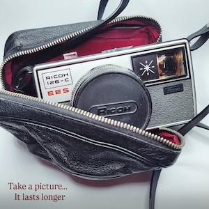Peut inclure: Un appareil photo Ricoh 126-C EES vintage avec un bouchon d'objectif noir à l'intérieur d'un étui en cuir noir. L'appareil photo a un corps argenté et un flash. L'étui a un intérieur rouge. Le texte "Take a picture... It lasts longer" est en bas.