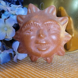 May include: A brown sun-shaped ornament with a smiling face, featuring pointed rays. The ornament is hanging by a string. Blue flowers and a gold-coloured surface are in the background. The ornament is on a yellow and white striped surface.