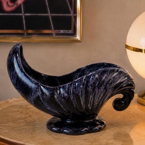 Peut inclure: Un bol décoratif en céramique avec un vernis marbré bleu marine profond et blanc. Le récipient présente une forme allongée et incurvée avec un bord évasé en forme de coquille et une extrémité en volute, reposant sur un socle circulaire festonné.