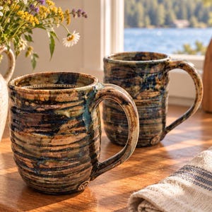 May include: Two ceramic mugs featuring a ribbed, textured surface with a variegated glaze in shades of deep blue, earthy brown, and tan. Each mug has a sturdy handle and a cylindrical body, showcasing a handcrafted appearance with a glossy finish.