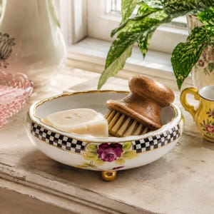 Peut inclure: Un porte-savon en céramique blanche avec une bordure à carreaux noirs et blancs, une bordure dorée et un motif floral. Il contient un pain de savon et une brosse en bois. Le plat repose sur trois pieds dorés.