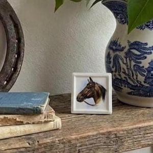 May include: A small square white ceramic tile featuring a detailed illustration of a brown horse head in profile. The horse is depicted with a dark mane and a bridle. The tile is positioned upright, showcasing the artwork clearly on its flat surface.