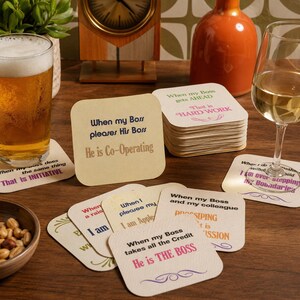 May include: A selection of coasters with witty office-themed sayings in various colours. The coasters are square and irregular in shape. A glass of beer and a glass of white wine are also visible on the wooden table.