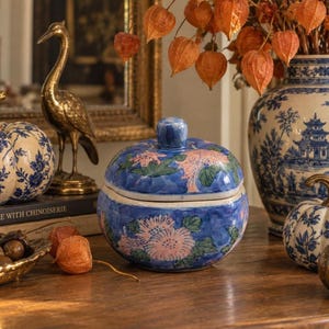 Peut inclure: Un pot en céramique rond avec couvercle, présentant un fond bleu orné de motifs floraux roses peints à la main et de feuilles vertes. Le pot possède un couvercle bombé assorti avec une petite poignée, affichant un design décoratif.